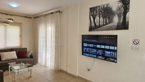 A living room with a sofa, a glass coffee table, a TV mounted on the wall displaying streaming apps, and a large framed picture of a tree-lined path. - 123 AN - Stay Nissi House (Ayia Napa)