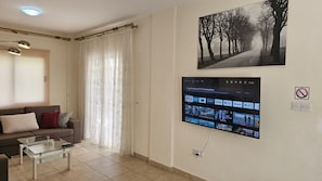 Un salón con sofá, una mesa de centro de vidrio, un televisor en la pared mostrando aplicaciones de transmisión y un cuadro grande con un camino bordeado de árboles.