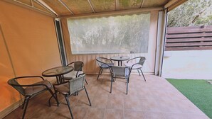 Un patio cubierto con cuatro sillas de mimbre y dos mesas redondas, una de ellas con un cuenco de metal. El espacio está delimitado por un muro de madera y una puerta de cristal, con vistas a un césped verde y árboles.
