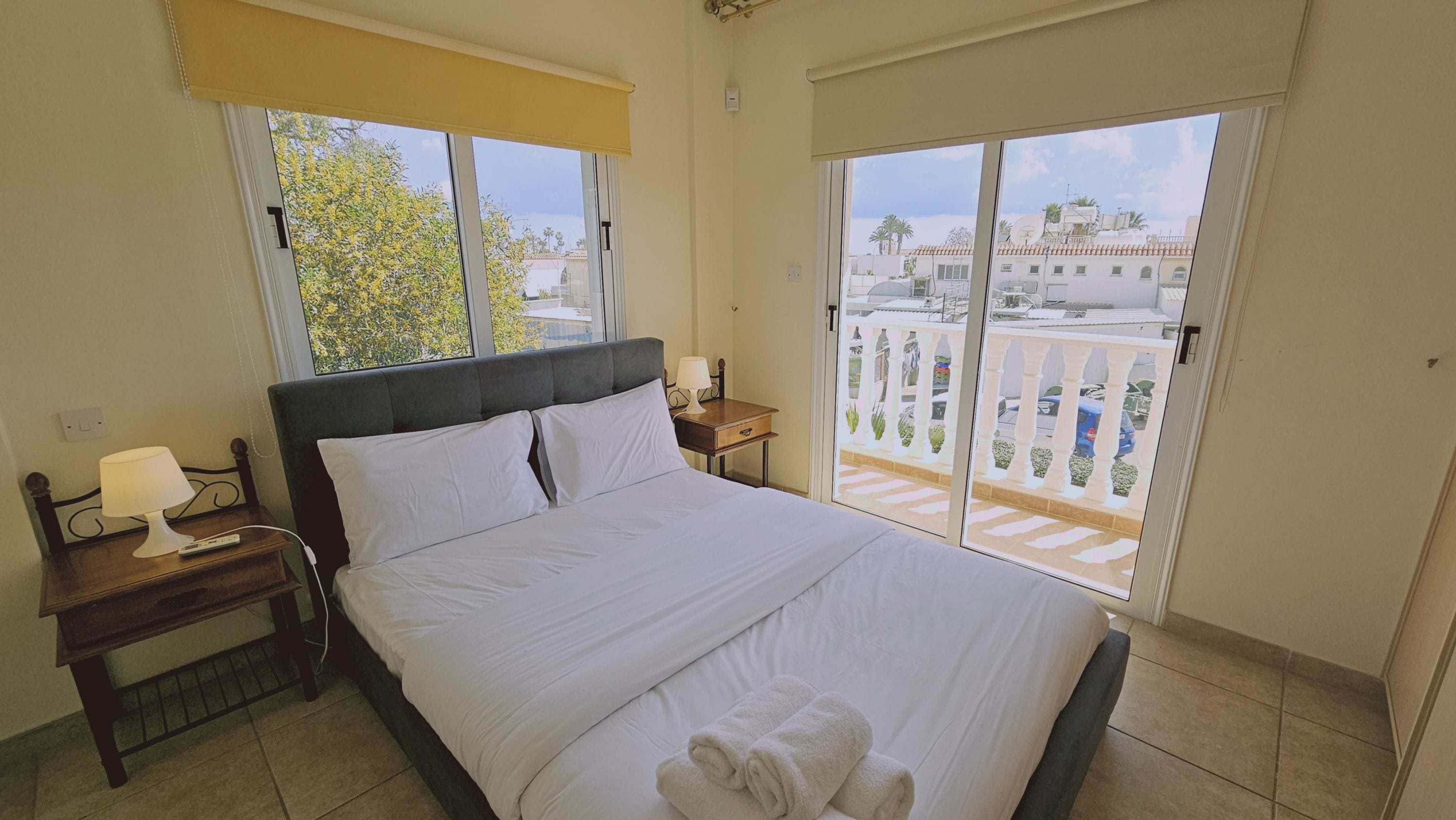 Una cama bien tendida con sábanas blancas, una mesita de noche con lámpara, y vistas a un balcón con barandilla y vegetación.
