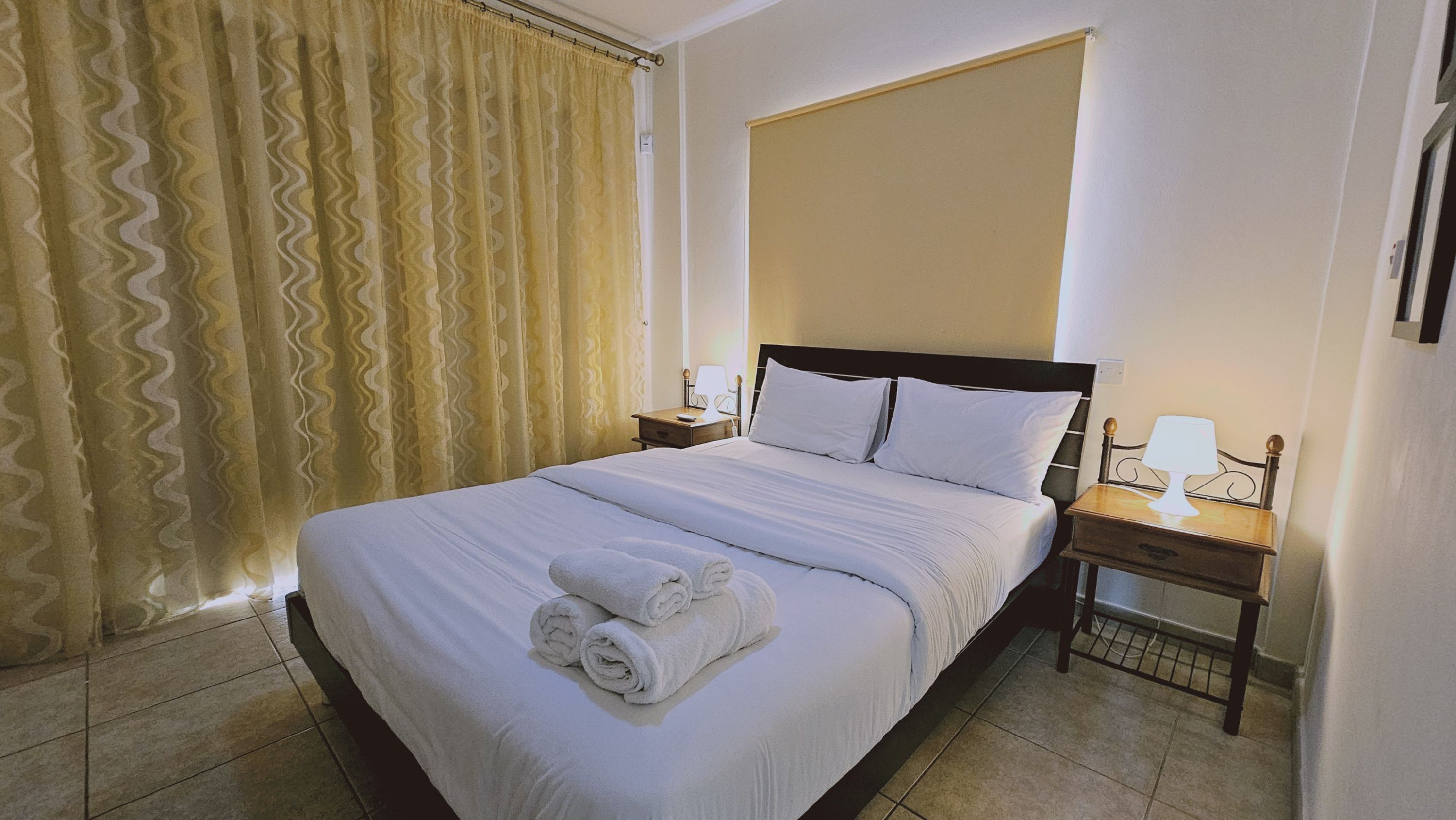 A neatly made bed with white linens and rolled towels, a bedside table with a lamp, and a window with patterned curtains.