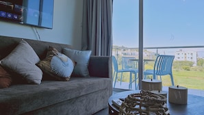 A modern living room with a grey sofa, a flat-screen TV mounted on the wall, and a large window offering a view of the outdoors. - 30 PRO - Stay White Coral Studio (Paralimni)