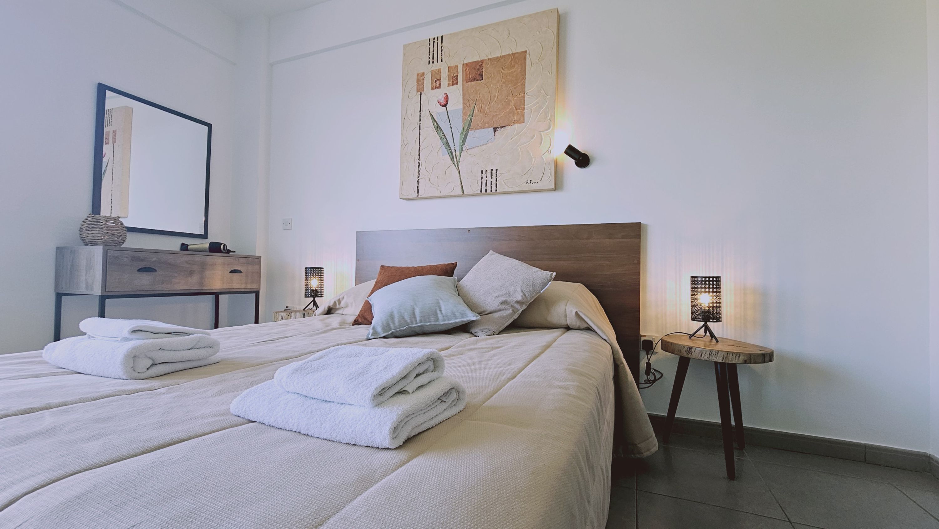 Una cama bien hecha con toallas dobladas, un cabecero de madera y dos lámparas de noche.