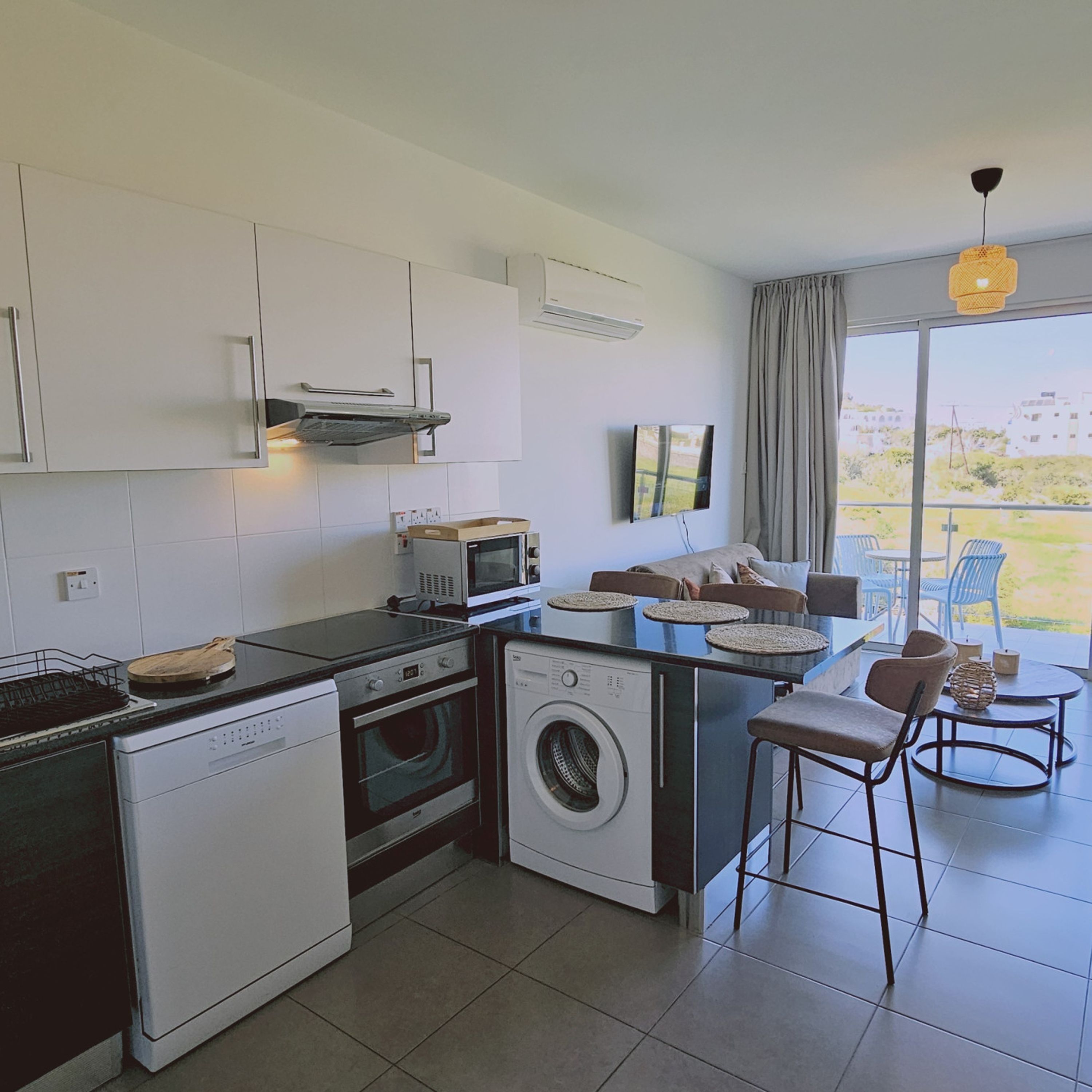 Cozinha moderna equipada com lava-louças, forno, micro-ondas e máquina de lavar roupa. Possui área de jantar com mesa e cadeiras, e uma grande janela com vista para o exterior.