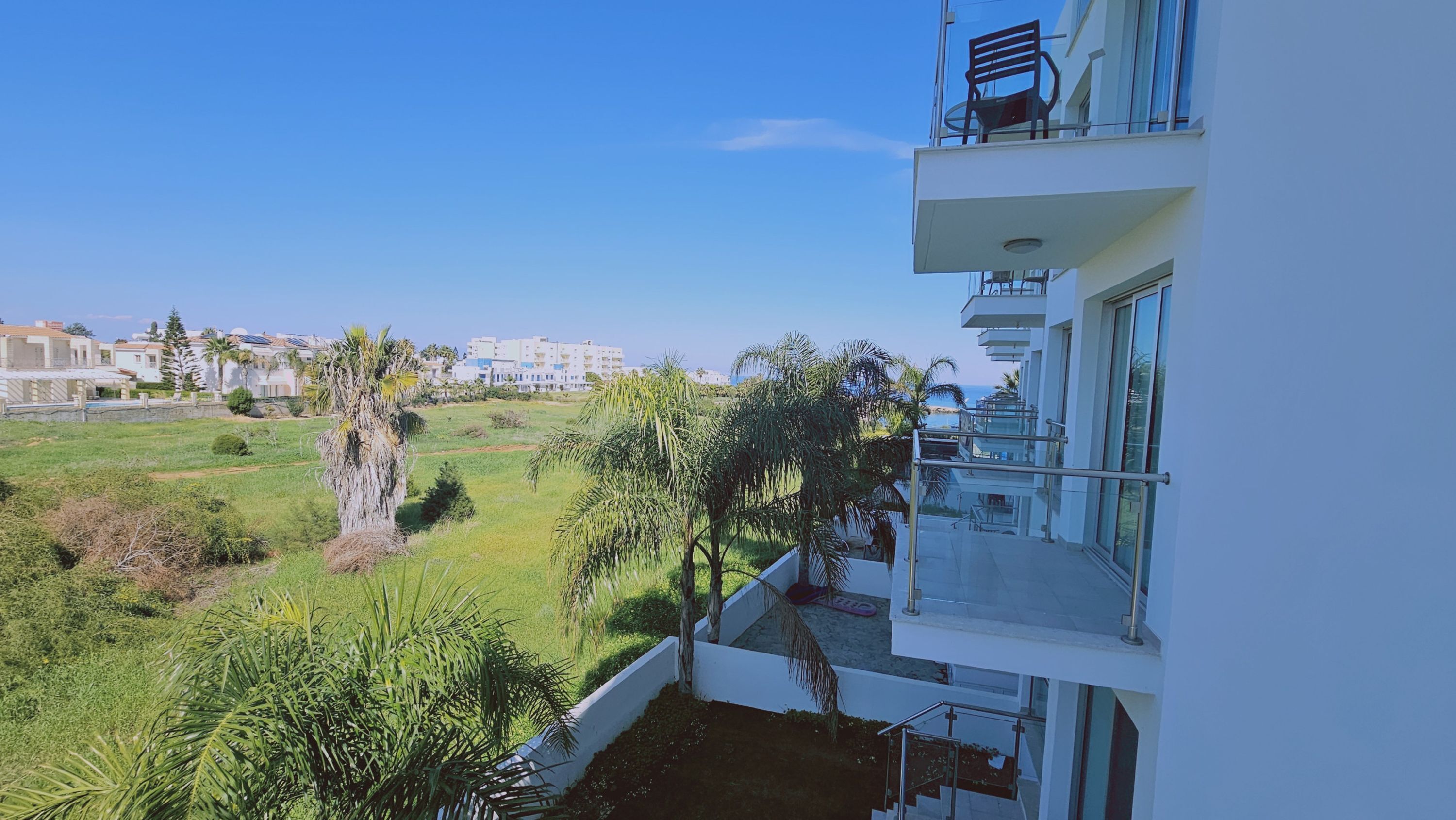 A modern building with a balcony overlooking a grassy area with palm trees and a view of the sea.