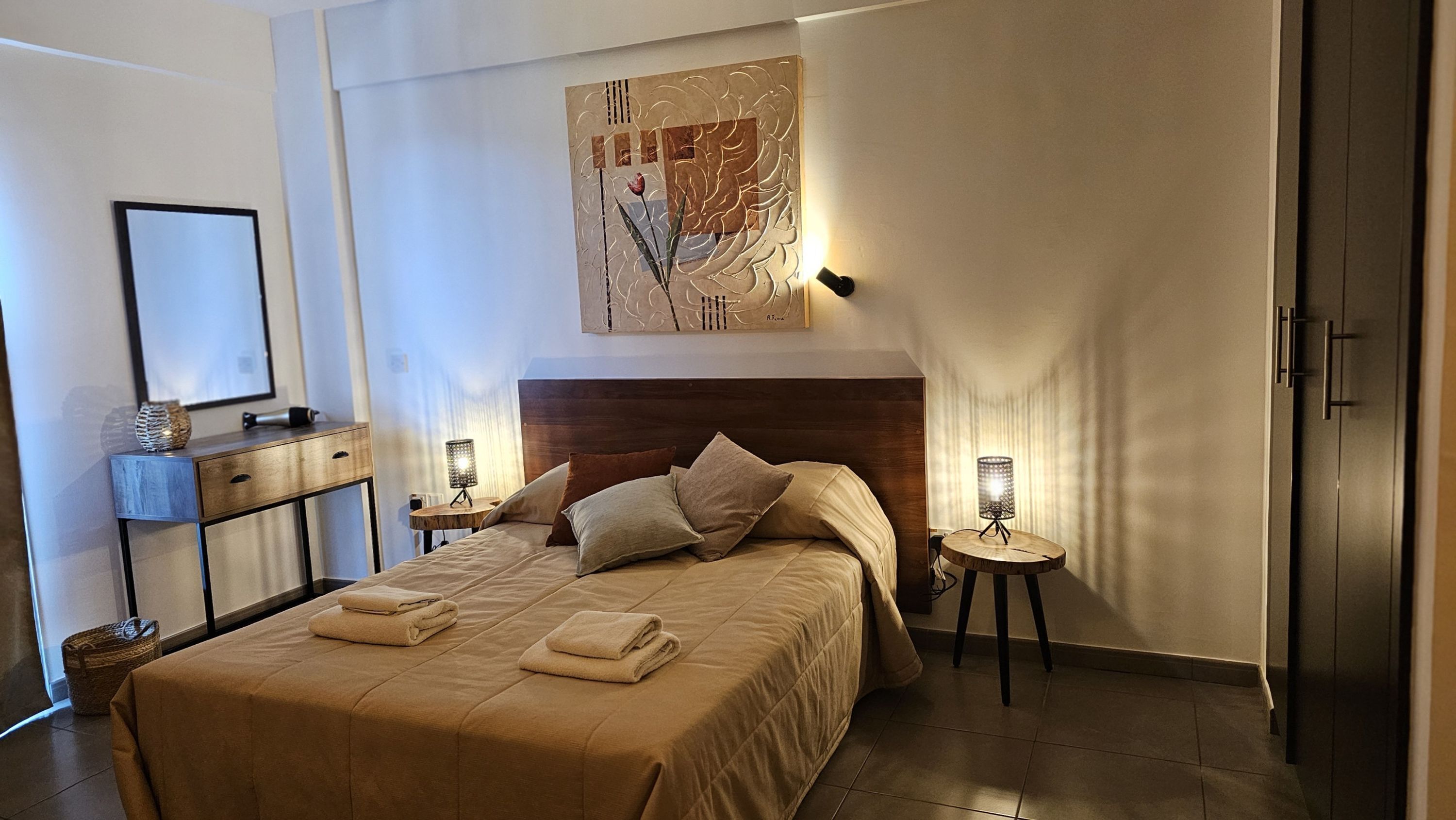 A modern bedroom with a large bed, two bedside tables with lamps, a wooden headboard, and a framed artwork above the bed.