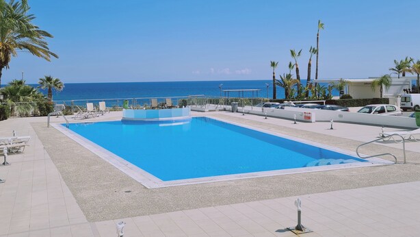 A rectangular swimming pool with a clear blue water, surrounded by a tiled deck, with palm trees and a view of the ocean. - 30 PRO - Stay White Coral Studio (Paralimni)