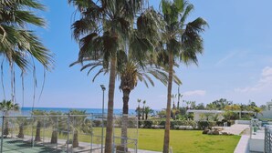 A fenced outdoor area with palm trees, a tennis court, and a view of the sea. - 30 PRO - Stay White Coral Studio (Paralimni)