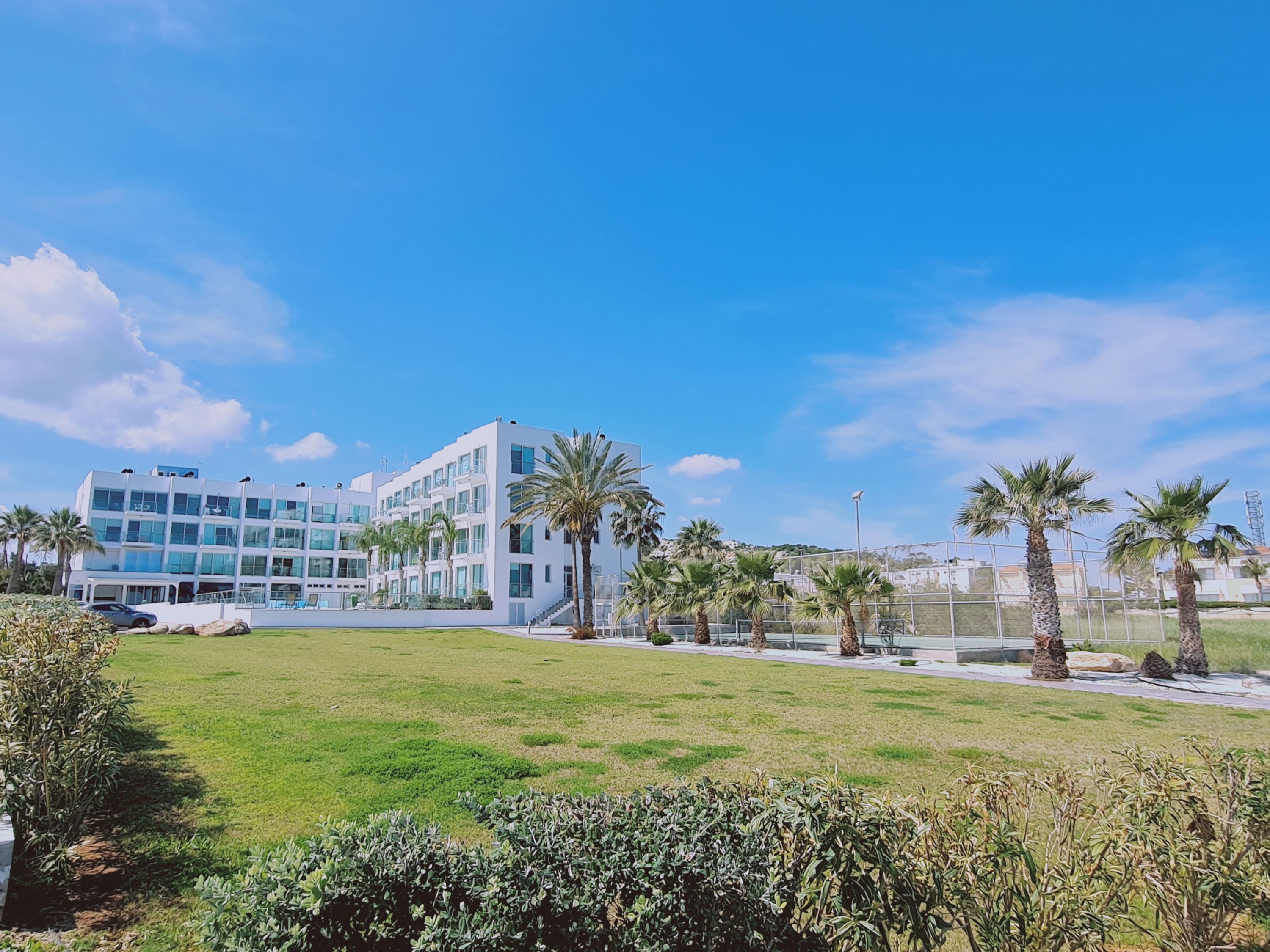 A modern building with multiple floors and large windows, surrounded by a well-maintained grassy area with palm trees.