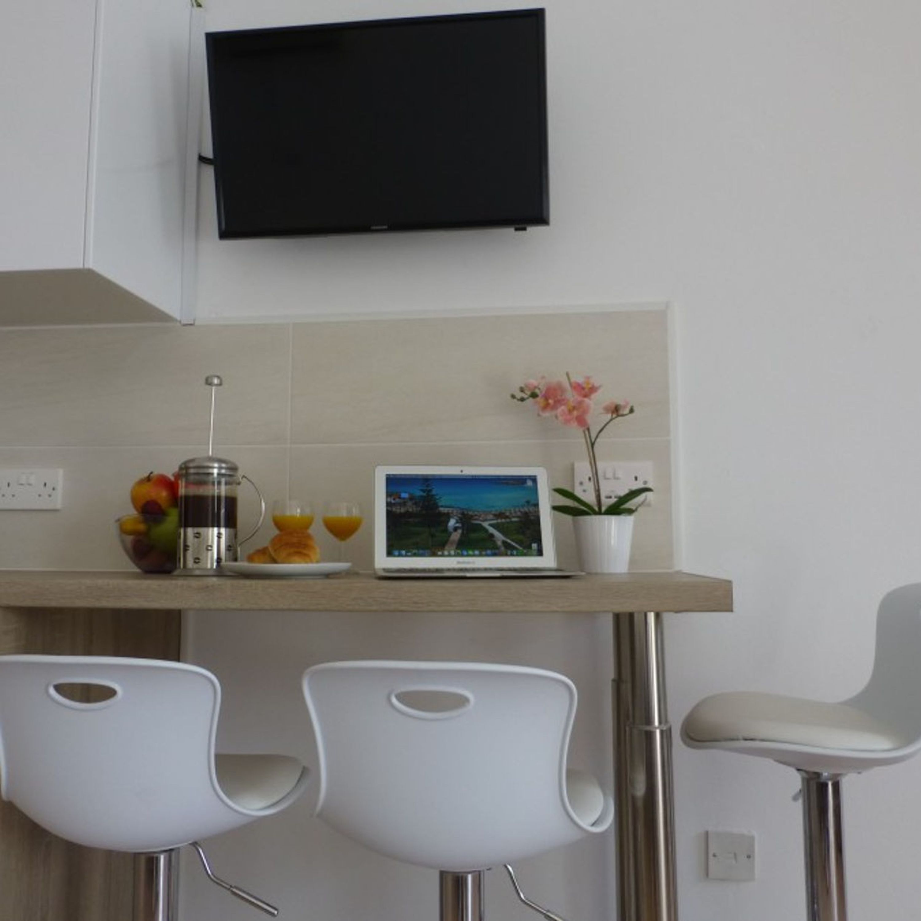 A modern kitchen with a flat-screen TV mounted above a wooden countertop. There are two white chairs, a laptop, a fruit bowl, and a coffee maker on the counter.