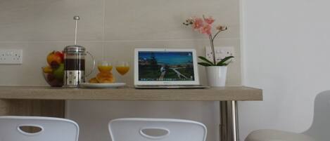 A modern kitchen with a flat-screen TV mounted above a wooden countertop. There are two white chairs, a laptop, a fruit bowl, and a coffee maker on the counter.