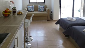 A compact hotel room with a kitchenette, a sofa with patterned cushions, a bed with a dark cover, and a large window. - 112 AN - Stay Paradise Sands Studio (Ayia Napa)