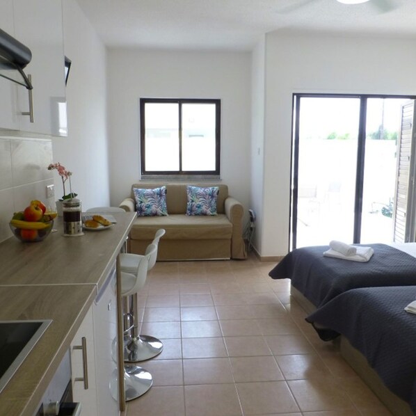 A compact hotel room with a kitchenette, a sofa with patterned cushions, a bed with a dark cover, and a large window.