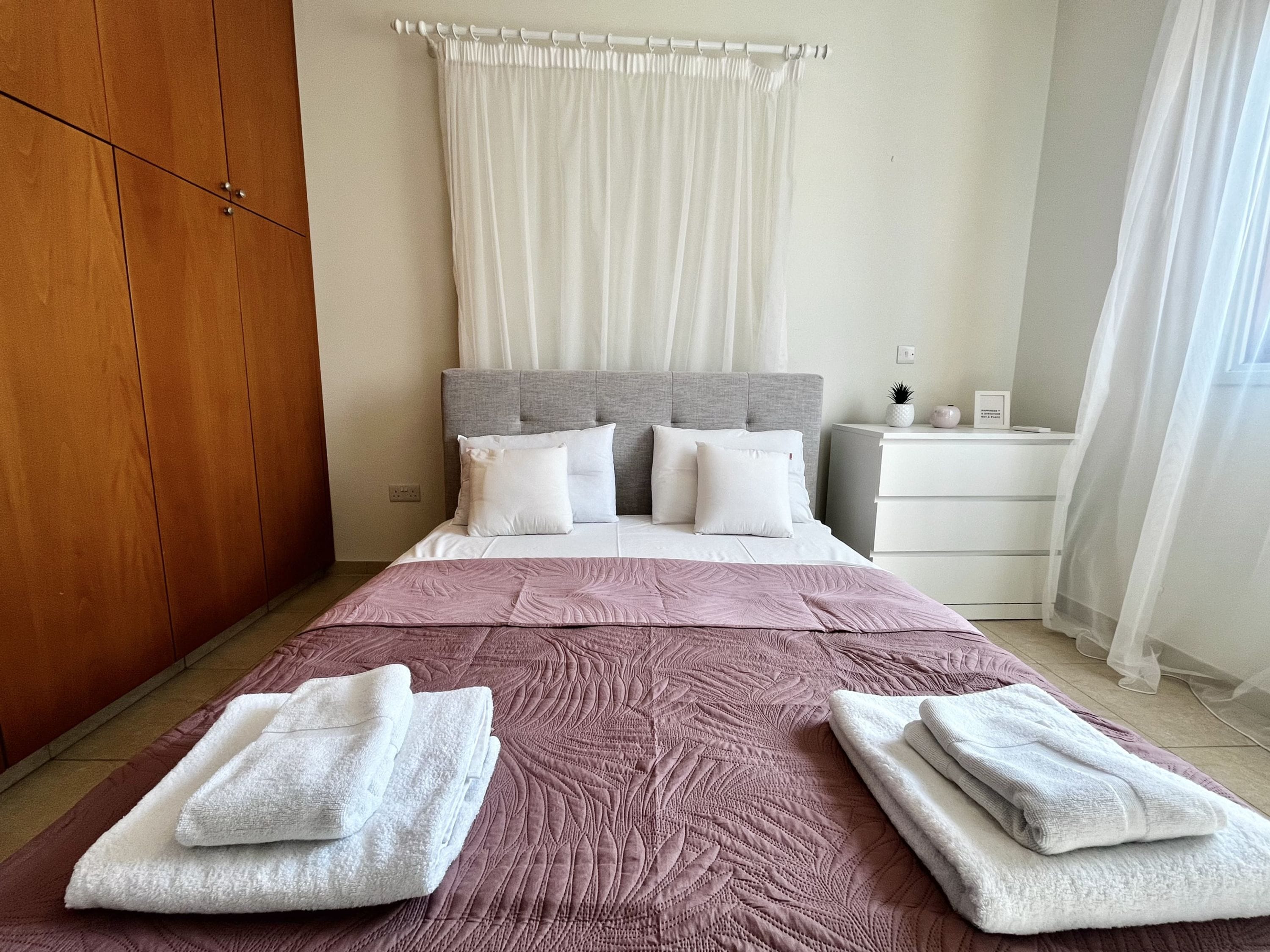 A neatly made bed with a pink quilt, a wooden wardrobe, and a white bedside table with a lamp.