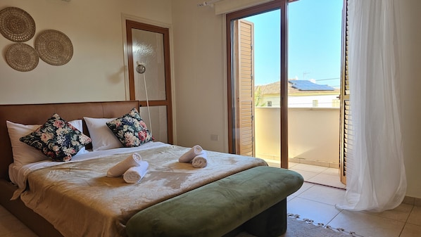 A hotel room with a bed, pillows, a bedside table, a window with blinds, and a view of buildings outside.