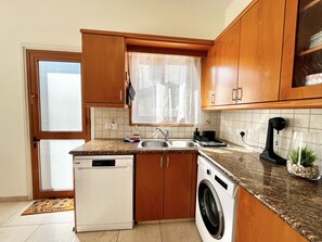 A compact kitchen with wooden cabinets, a dishwasher, a washing machine, and a window with a sheer curtain. - 111 VOR - Stay Tranquility Cove (Oroklini)