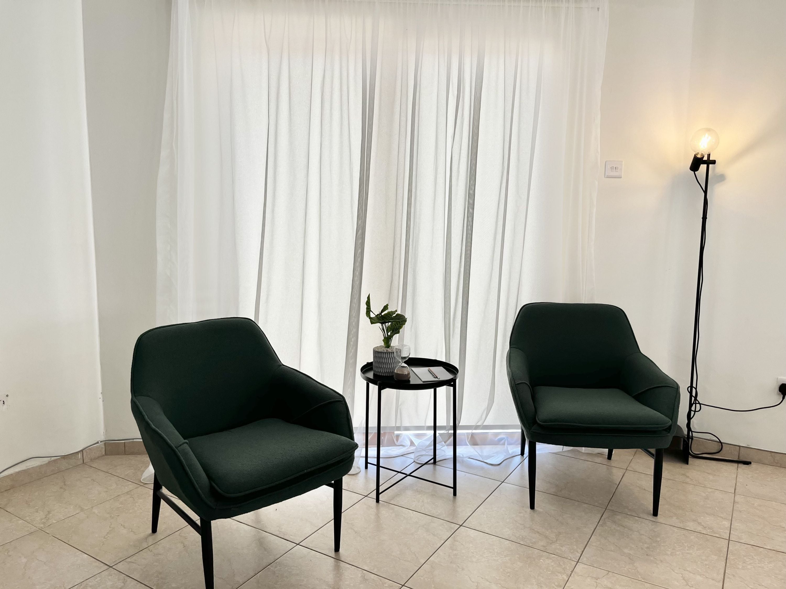 A room with two dark green chairs, a small table with a plant, and a floor lamp.