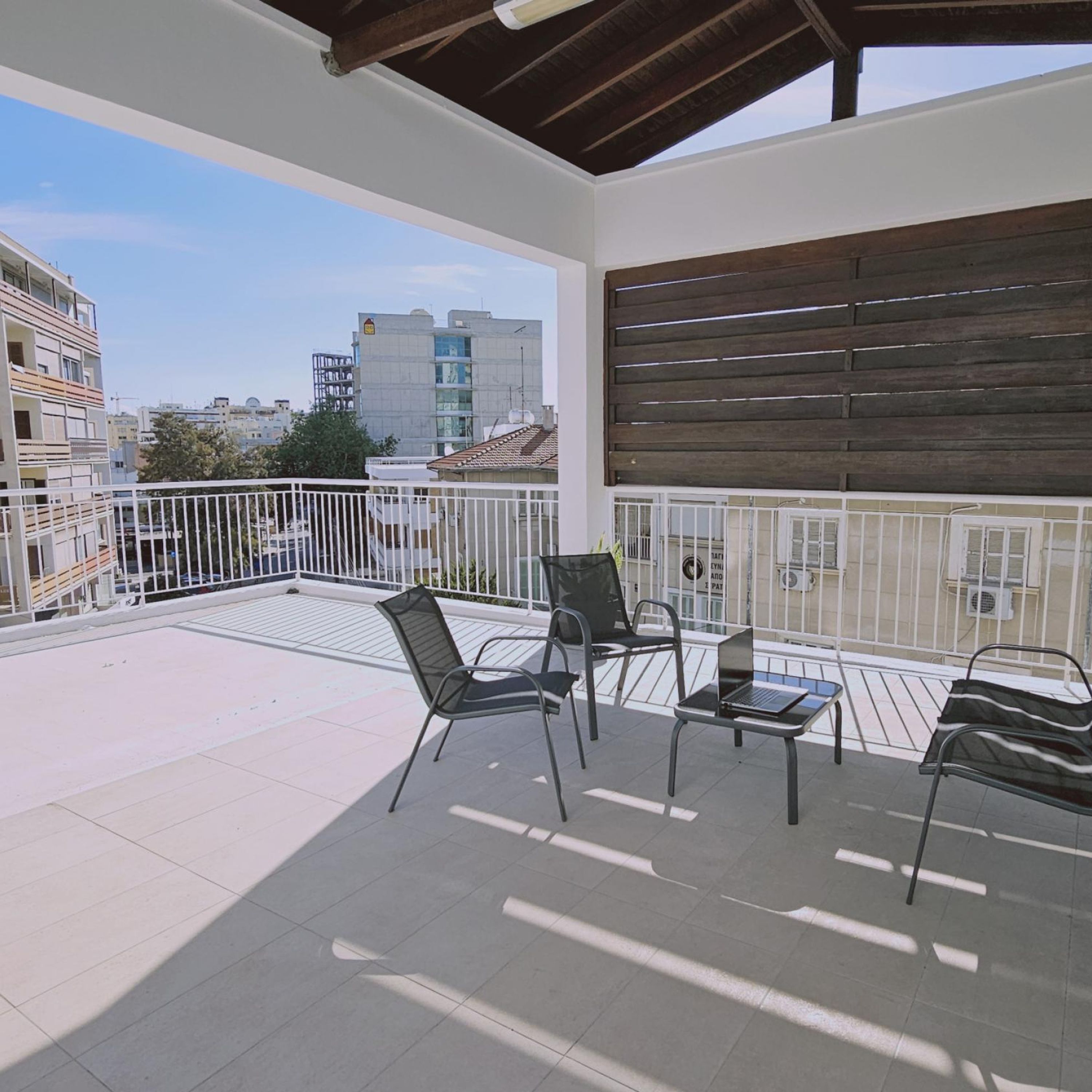 A balcony with a view of city buildings, metal chairs, and a table with a laptop.