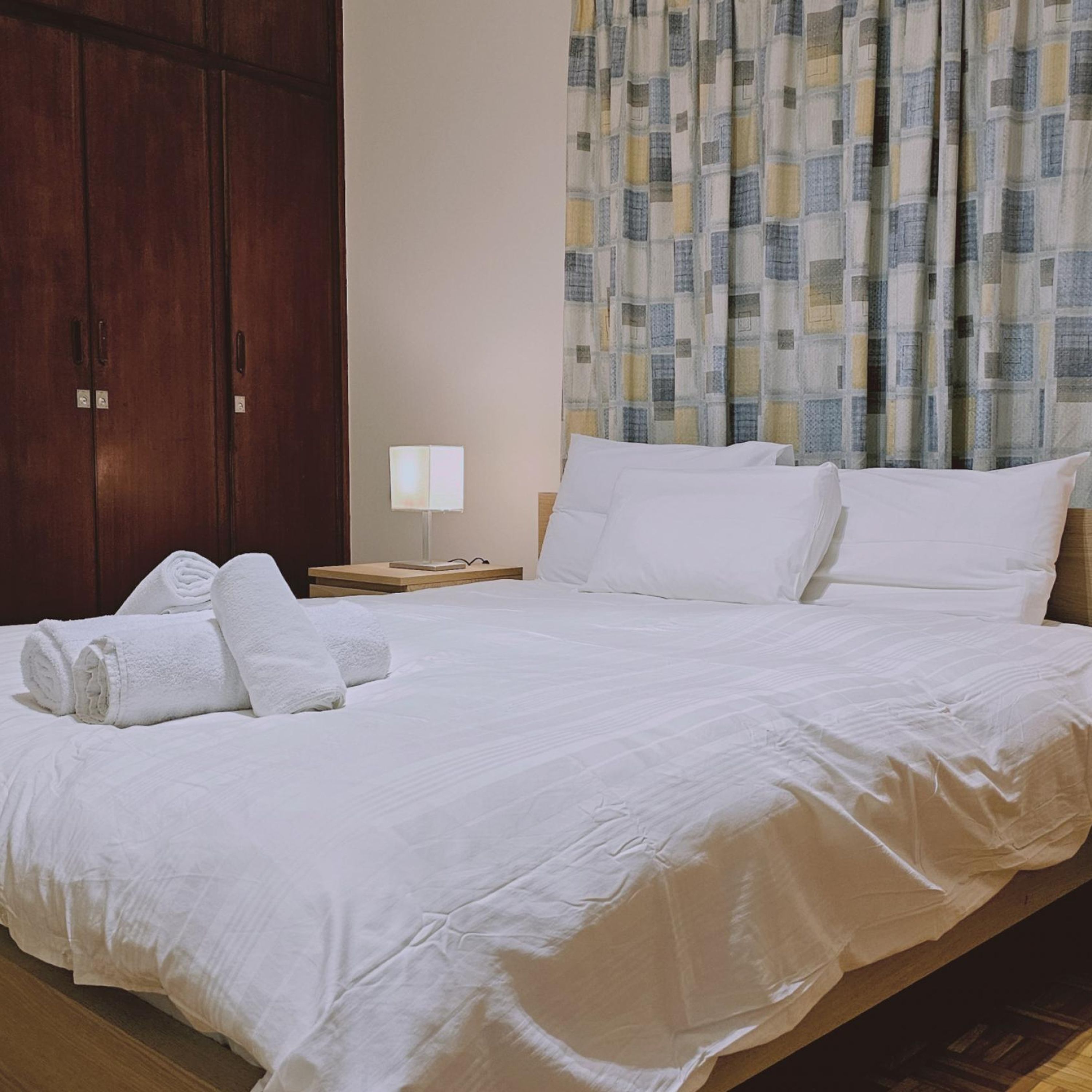 A neatly made bed with white linens, a wooden wardrobe, and a patterned curtain.