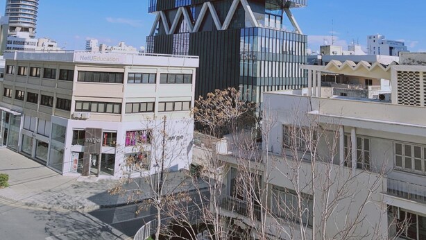 Un bâtiment moderne au design original, caractérisé par une forme en zigzag, entouré d’autres immeubles et surplombé d’un ciel bleu azur.