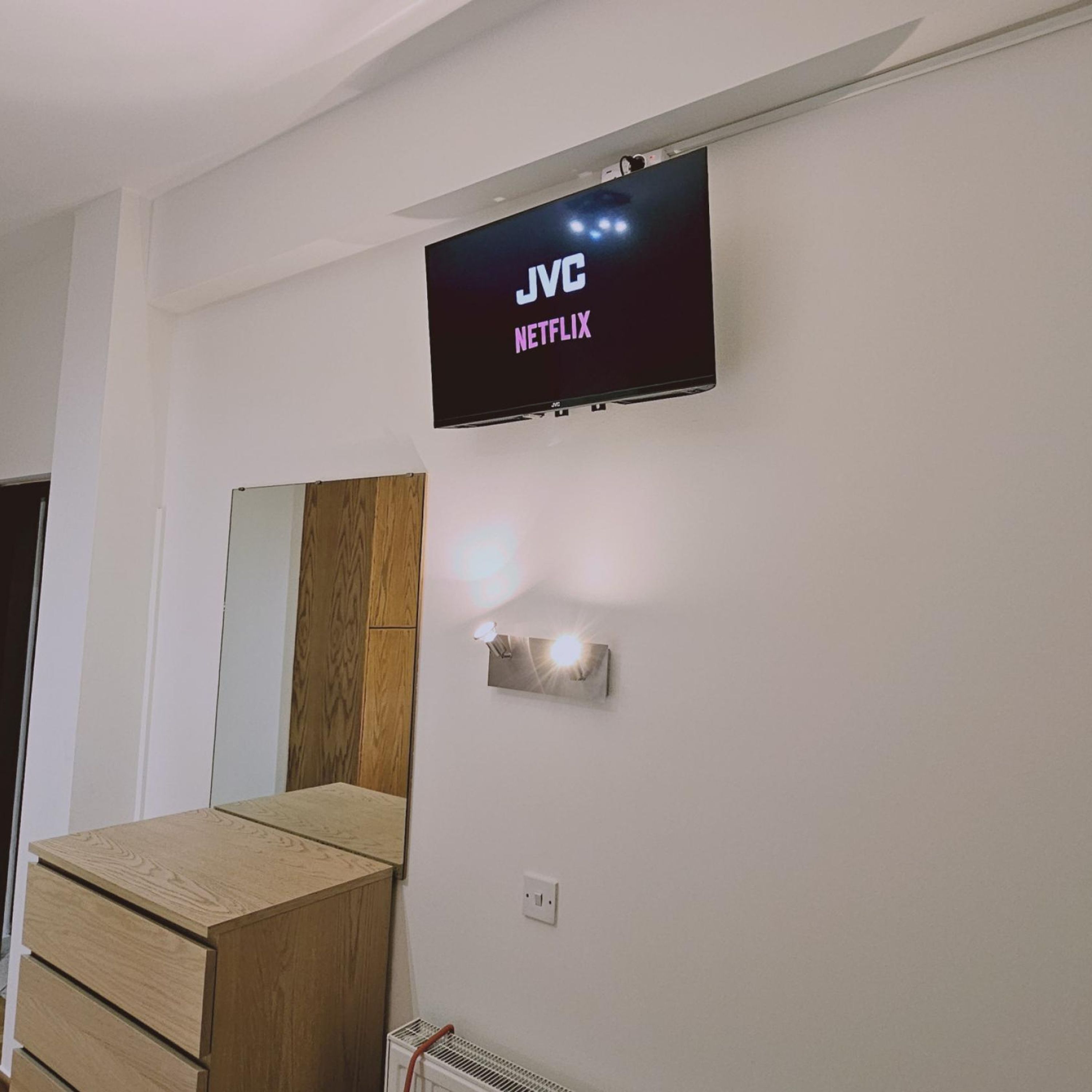 Une pièce avec un téléviseur fixé au mur qui affiche « JVC Netflix », un meuble en bois et un luminaire fixé au mur.