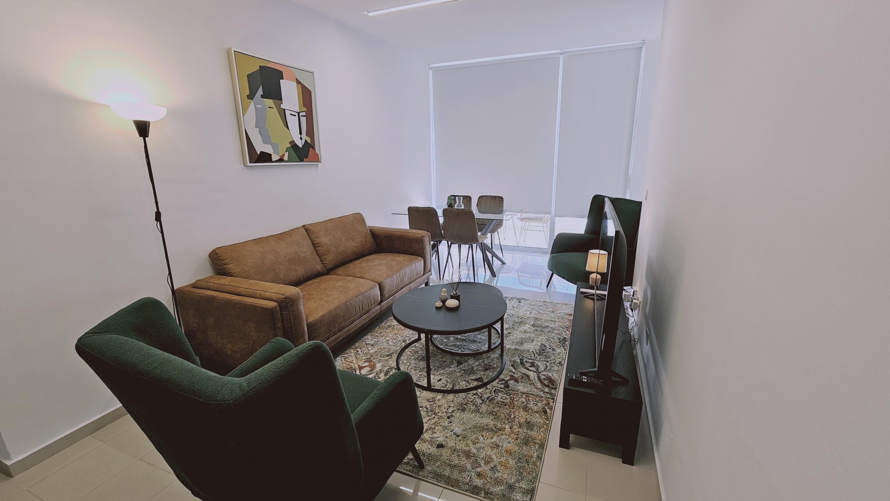 A modern living room with a brown sofa, green armchair, and a round coffee table. There is a painting on the wall and a TV stand with a lamp.