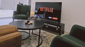 A modern living room with a flat-screen TV displaying the Netflix logo, a green armchair, a brown sofa, and a coffee table with decorative items.