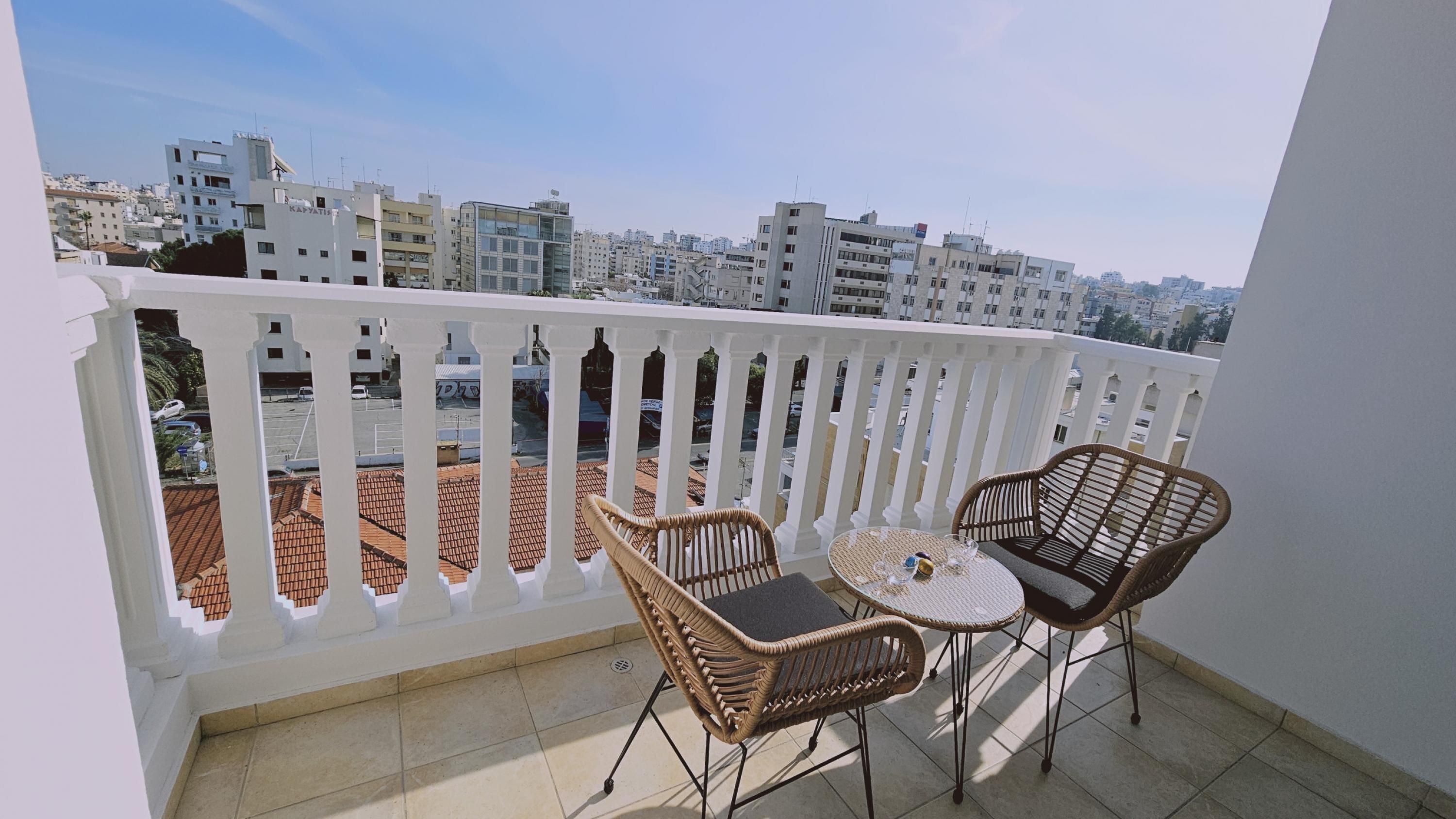 A balcony with two wicker chairs and a small round table, overlooking a cityscape with terracotta-tiled roofs and modern buildings.