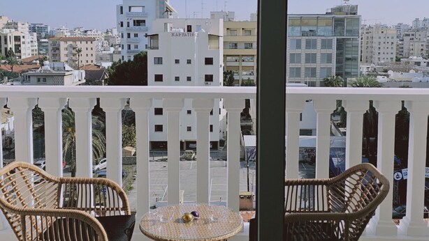 A balcony with wicker chairs and a small round table, overlooking a cityscape with various buildings.