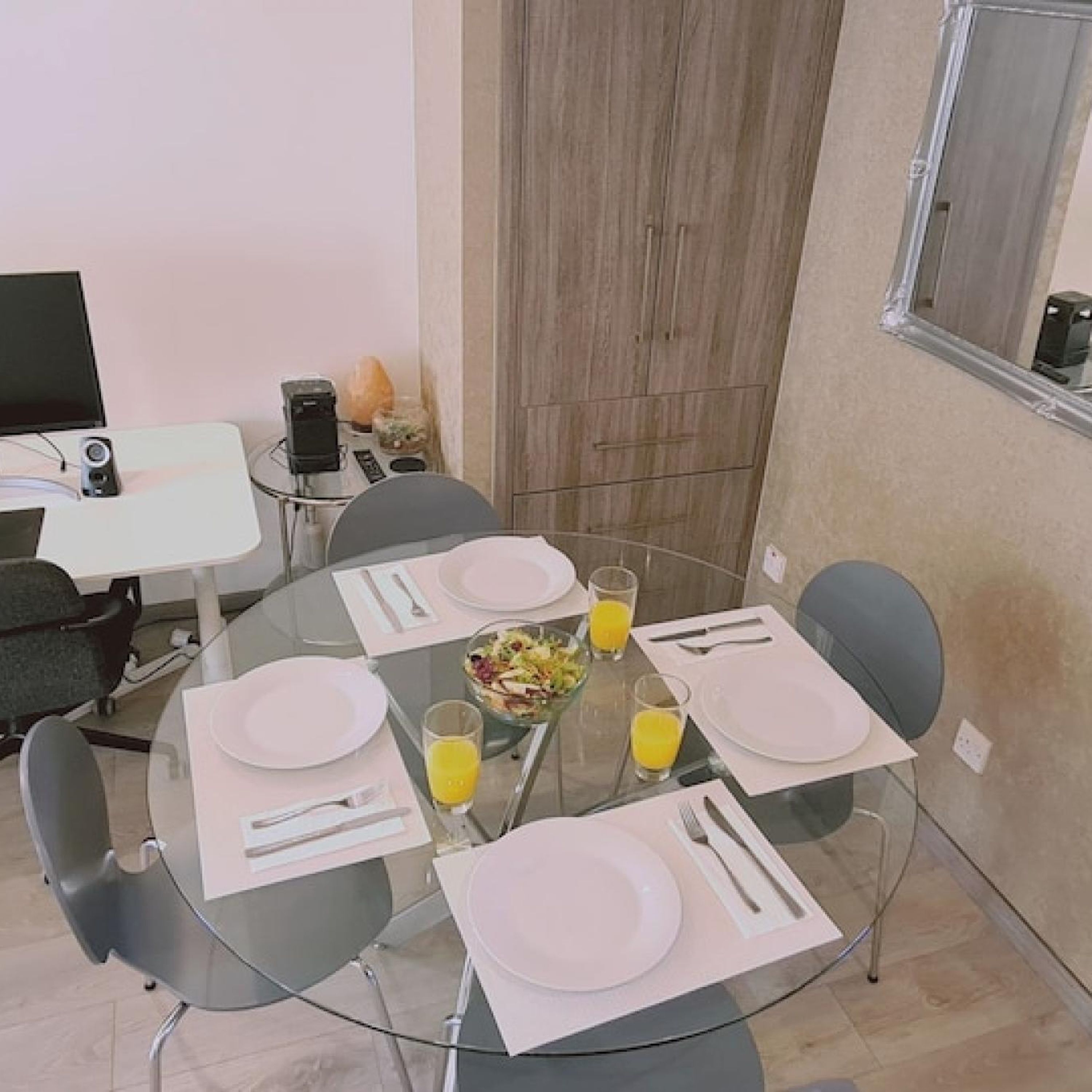 Una zona d'esmorzar moderna amb una taula rodona de vidre preparada per a quatre persones, amb plats, coberts i verres de suc d'aranja.