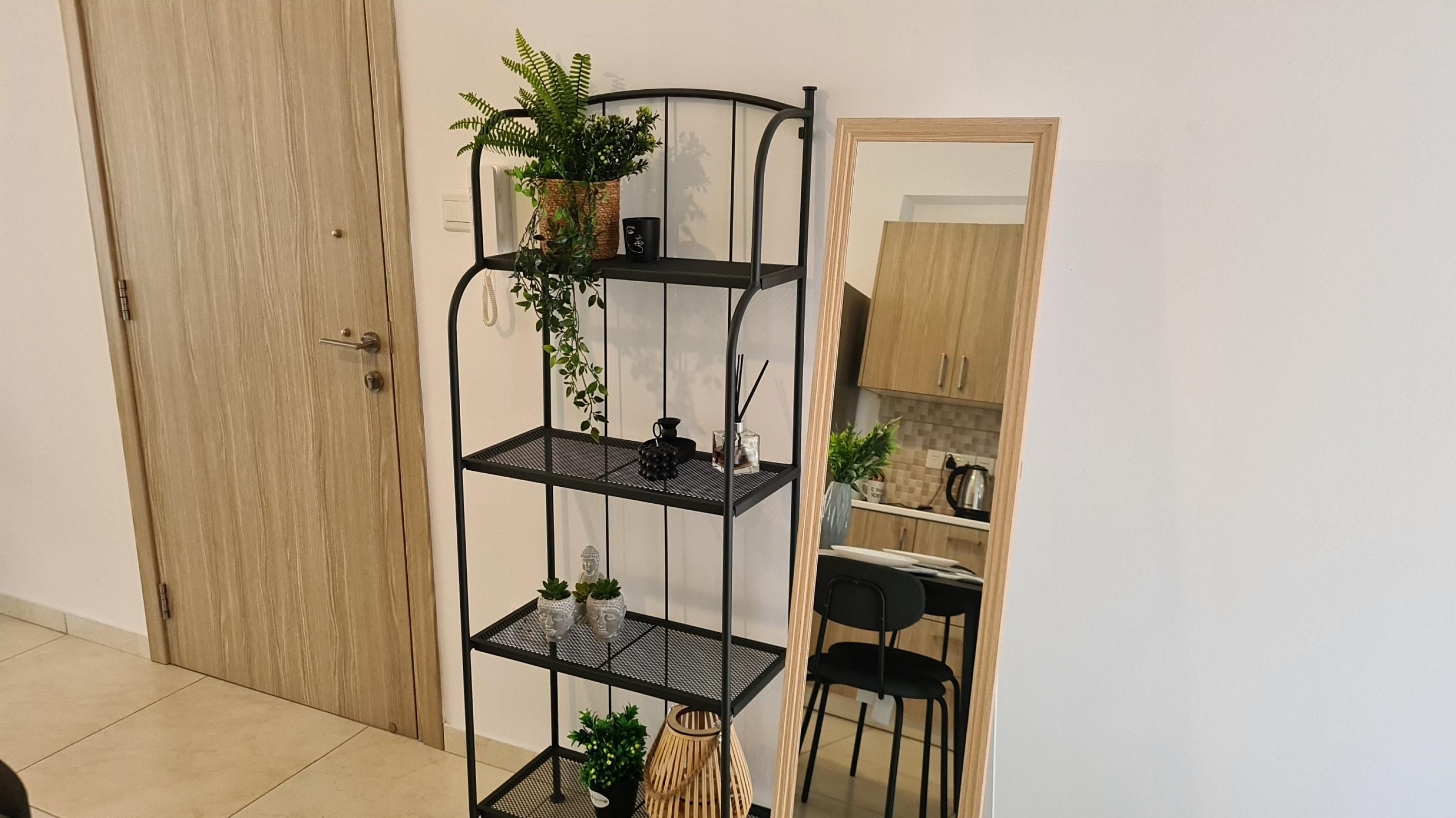 Estantería metálica negra de tres niveles con plantas y objetos decorativos, una puerta de madera y una cocina con armarios y una silla.