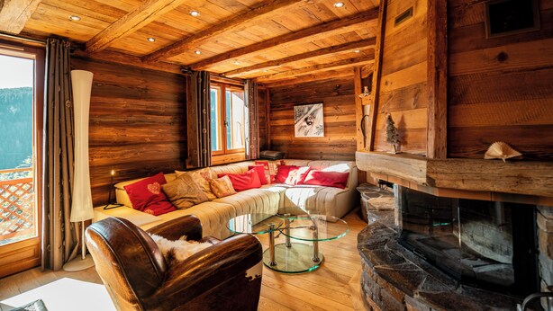 Ein gemütliches Blockhaus-Interieur mit Holzbalkendecke, Kamin aus Stein, Ledersofa und Glastisch.