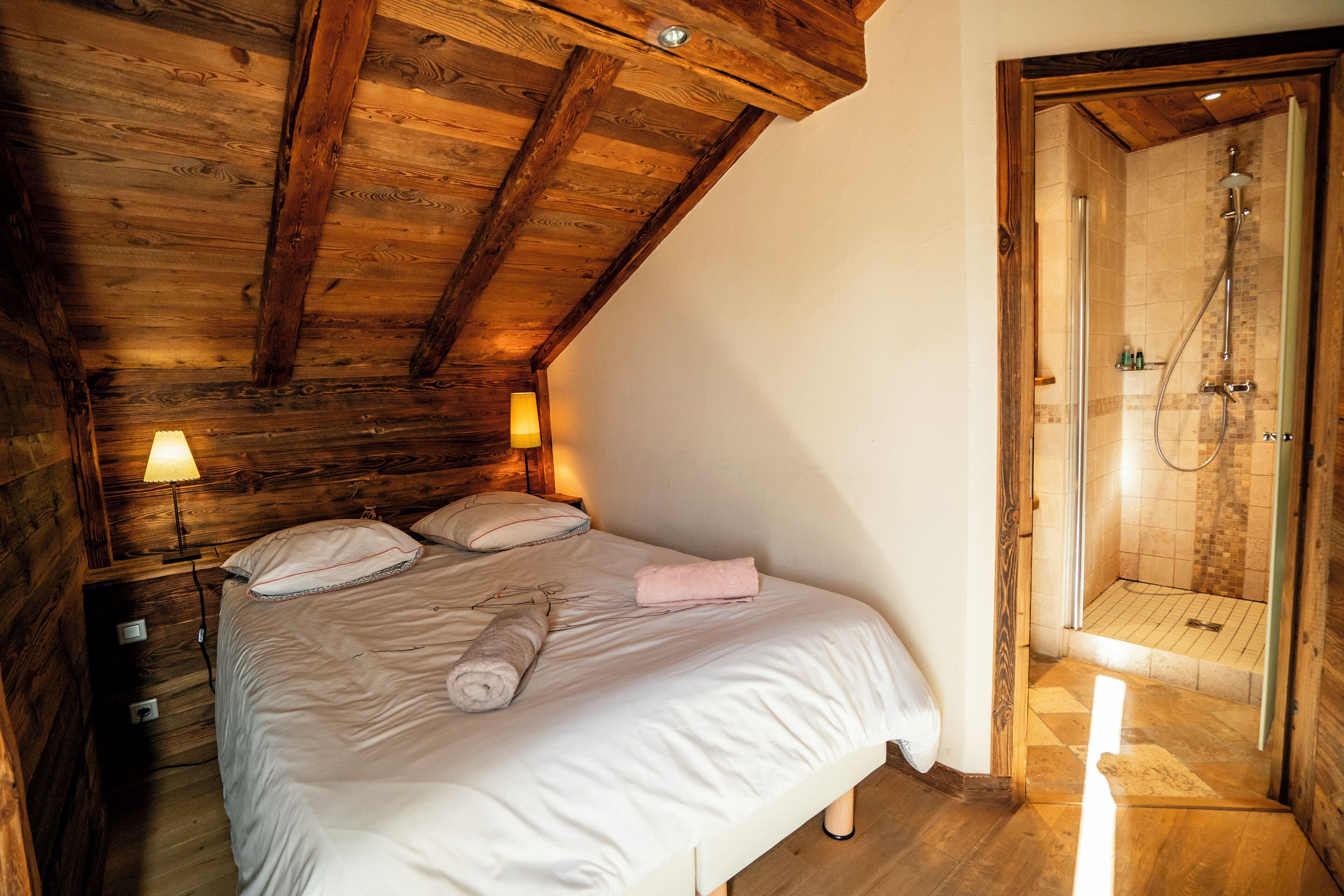 Een slaapkamer met een houten plafond, een bed met witte lakens, een roze handdoek, en een houten deur die leidt naar een douchecel.