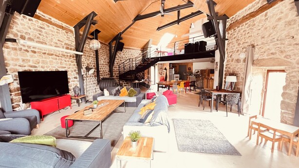 A spacious living area with a stone wall, wooden ceiling, and a large TV. There are sofas, a coffee table, and a dining table with chairs. - Gite de Groupe Lozère Aubrac (Peyre en Aubrac)