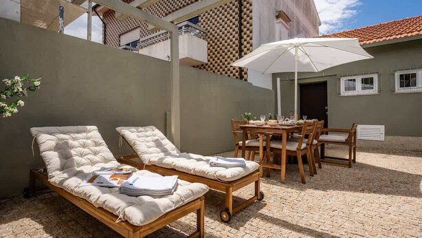 Eine sonnendurchflutete Terrasse im Freien mit zwei Liegestühlen, einem Holztisch und Stühlen sowie einem weißen Sonnenschirm.