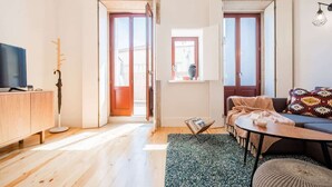 Una sala de estar moderna con un sofá, una mesa de centro y un televisor sobre un mueble de madera. Se ve una ventana con vistas al exterior.