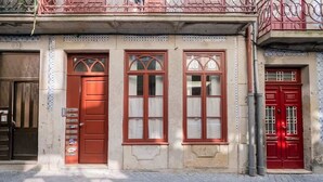 Fachada de un edificio con tres puertas rojas, ventanas y barandilla metálica para el balcón, además de pavimento de baldosas.