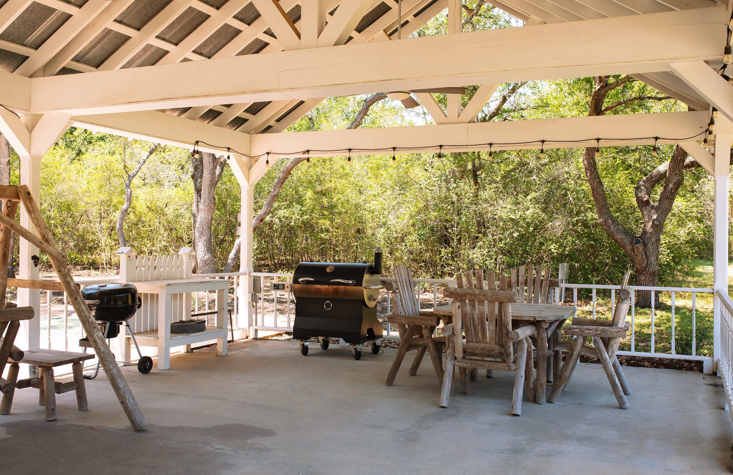 An outdoor covered patio with a grill, wooden chairs, and a white fence.