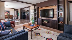 A modern living room with a large TV, dark wood cabinets, a dining area, and a kitchen in the background. - Wimberley | Hot Tub+Fire Pit+Pickleball | Sleeps 6 (Wimberley)