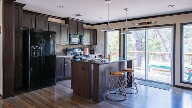 A modern kitchen with dark wood cabinets, a black refrigerator, a microwave, and a large island with stools. - Wimberley | Hot Tub+Fire Pit+Pickleball | Sleeps 6 (Wimberley)