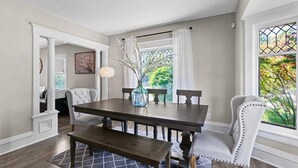Ein Esszimmer mit einem dunklen Holztisch, weiß gepolsterten Stühlen und einer Bank. Auf dem Tisch steht eine Vase mit Zweigen, und ein Fenster bietet Blick auf Grünflächen.