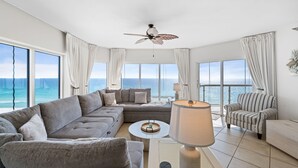 A modern living room with a large sofa, a striped armchair, and a coffee table. There is a ceiling fan and a view of the ocean through large windows. - Breathtaking Gulf Views! Community Pool! Steps to the Gulf! (Pensacola Beach)