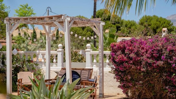 A wooden pergola with string lights, outdoor seating, and a lush garden with purple flowers. - 5-bed chalet in Albir, with swimming pool, AC, 20-min-walk to beach.  (l'Alfàs del Pi)