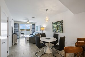 Un espace repas moderne avec une table ronde et des chaises noires, un ventilateur de plafond et une vue sur l’extérieur grâce à de grandes fenêtres.