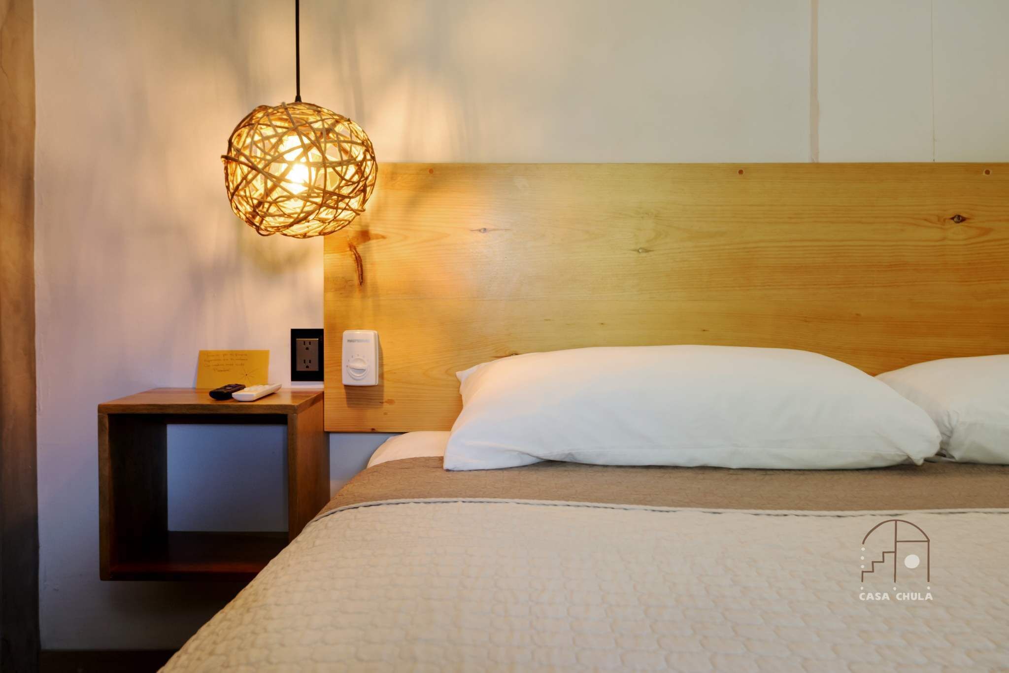 A neatly made bed with white pillows and a textured beige blanket. A unique woven light fixture hangs above the bed, and a small wooden nightstand with a remote control and a framed picture is to the left.