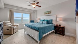 A bedroom with a bed, a recliner, a nightstand, a ceiling fan, and a window with a view of the ocean.