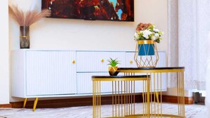 Un salon moderne avec un grand tableau représentant un feu, un meuble blanc, une table à cadre doré et un vase rempli de fleurs.
