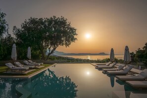 Un espace aménagé au bord de la piscine, avec des chaises longues et des parasols, offrant une vue dégagée sur le coucher de soleil sur l’eau, et un grand arbre qui procure de l’ombre.