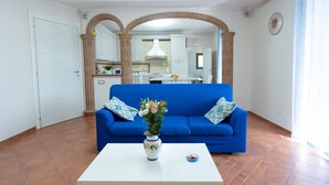 Un soggiorno con un divano blu, un tavolino da caffè bianco con un vaso, e una cucina sullo sfondo.