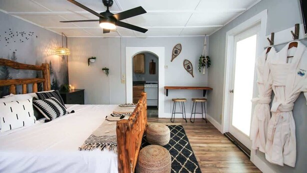 Ein Schlafzimmer mit einem Bett, einem Holz-Kopfende, Nachttischen, einem Deckenventilator, einem an der Wand montierten Fernseher und Blick in die Küche.